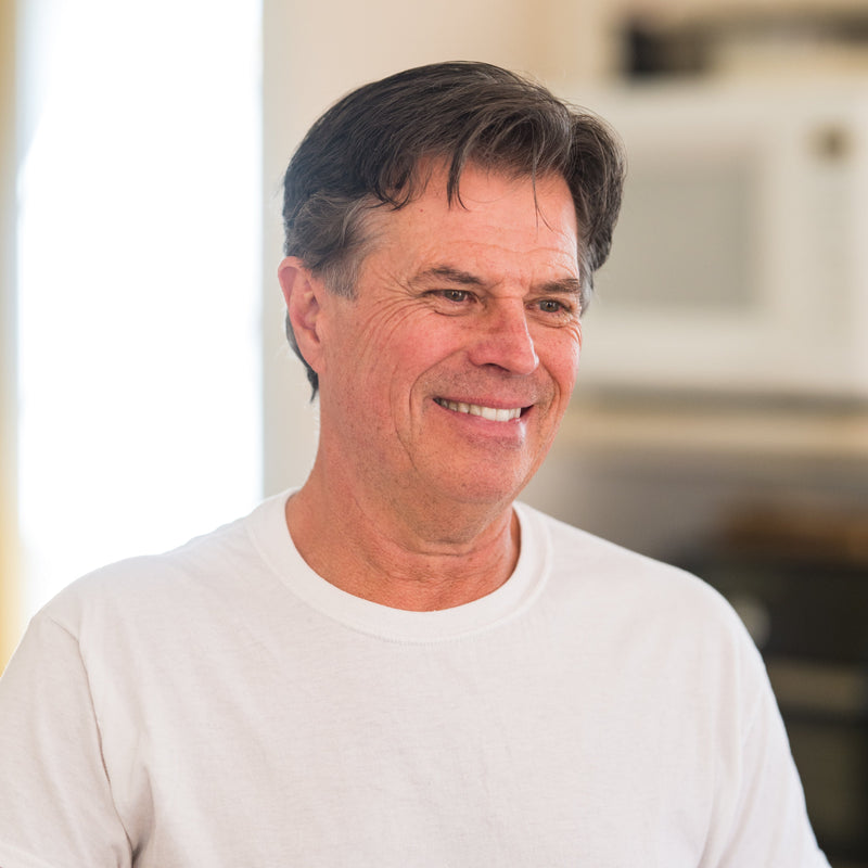 Image of Tim, owner of Tim's Serious Shake, wearing a white t-shirt in a blurred kitchen and smiling