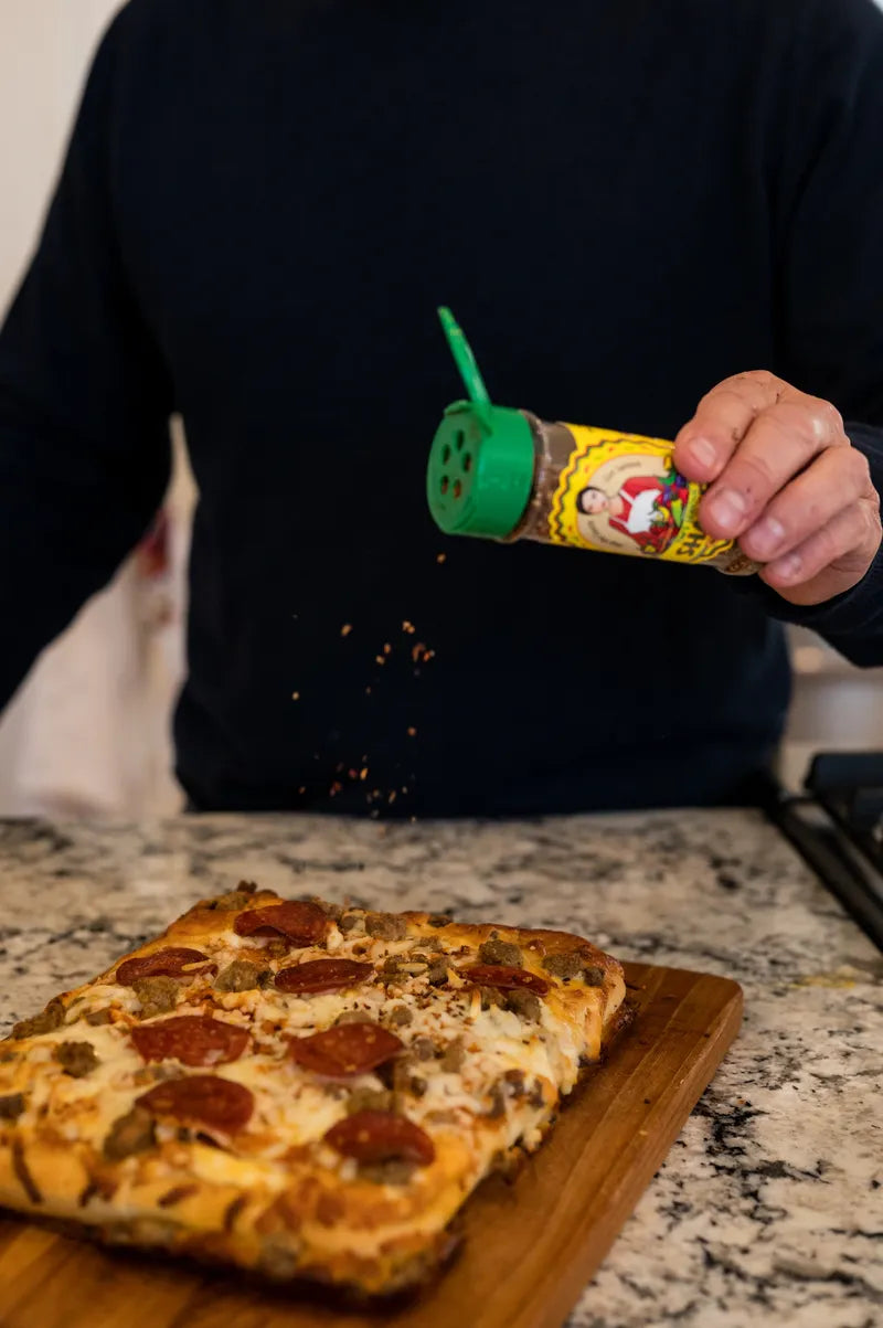 A person sprinkling seasoning from a bottle of Tim's Serious Shake onto a pizza on a wooden board.